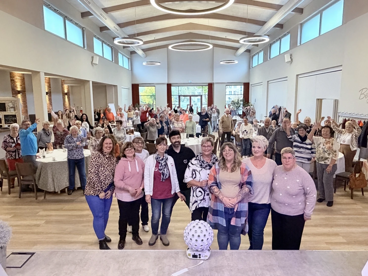Klientinnen, Klienten und das Team der Herzlichen Helfer feiern gemeinsam im Saal und winken für ein Gruppenfoto.