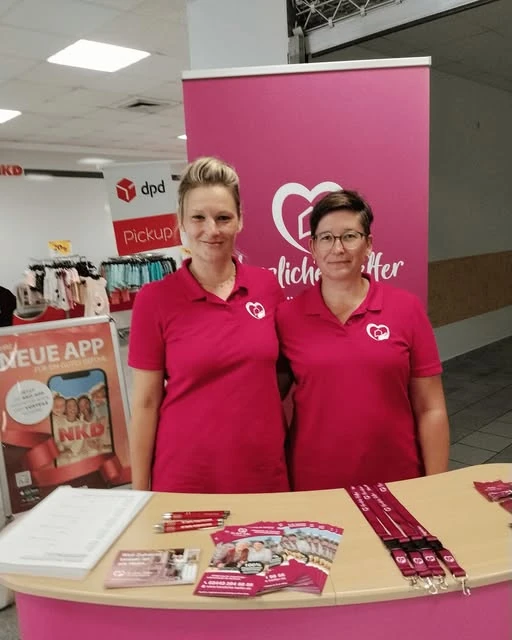 Zwei Teammitglieder der Herzlichen Helfer in weinroten Shirts an einem Infostand.