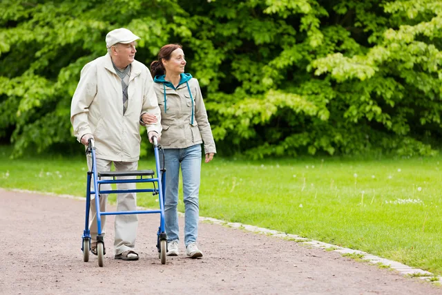 Ein älterer Mann geht mit einem Rollator und einer Begleitung auf einem Weg im Grünen.