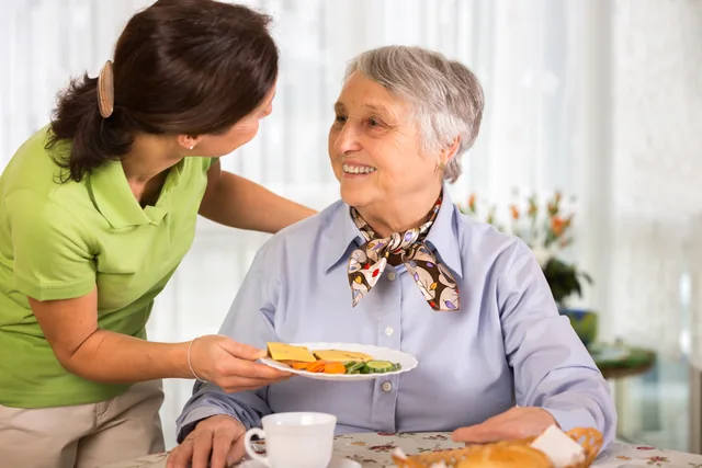 Eine Begleiterin bringt einer älteren Frau eine Mahlzeit an den Tisch.